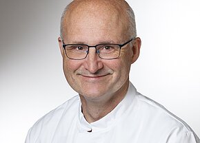 MU Dr. Jan Toman Ein lächelnder Mann mit Brille trägt einen weißen Laborkittel. Die Hintergrundfarbe ist hell, was ein professionelles Ambiente vermittelt. Der Mann strahlt Freundlichkeit und Vertrauen aus.