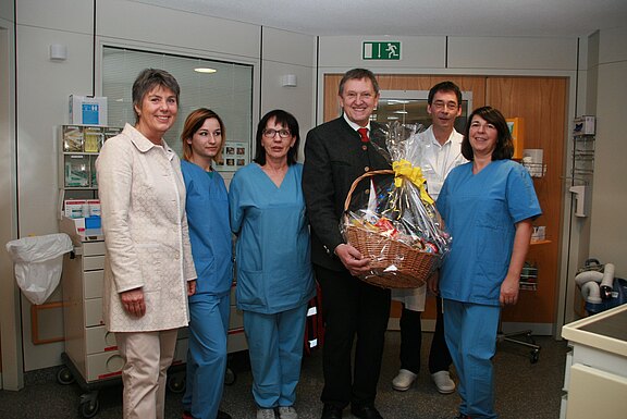 In einem Krankenhausflur steht ein Mann im Anzug mit einem großen, verpackten Geschenkkorb neben mehreren Pflegekräften in blauen Kitteln. Die Gruppe lächelt in die Kamera, als ob eine Spende oder ein Dankeschön übergeben wird.