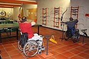 Zwei Personen im Rollstuhl üben in einer Halle Bogenschießen: Sie spannen Bögen und zielen auf Zielscheiben; an der Wand hängen weitere Bögen. Szene eines inklusiven, barrierefreien Sports.