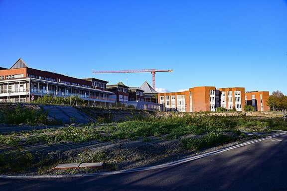 Neubaukomplex mit mehrstöckigen Backstein- und Glasgebäuden; ein roter Baukran ragt darüber. Im Vordergrund eine brachliegende, teils begrünte Fläche und eine Straße unter blauem Himmel.