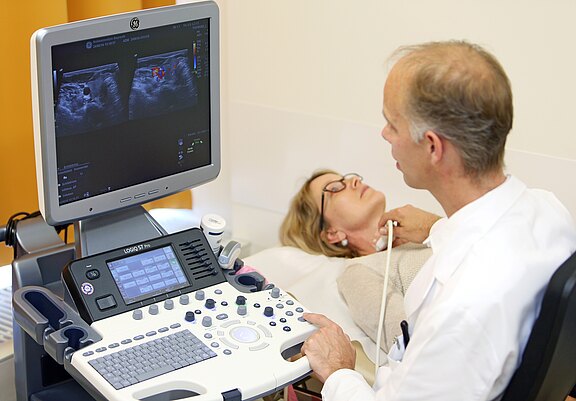 Ein Arzt führt einen Ultraschalluntersuchung bei einer Patientin durch. Der Monitor zeigt Ultraschallbilder, während der Arzt mit einem Sensor den Hals der Patientin untersucht. Die Patientin liegt entspannt auf einer Liege und schaut nach oben.