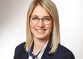 Corinna Greller Porträtfoto: lächelnde Frau mit schulterlangem blondem Haar und Brille, in dunkelblauem Blazer und weißer Bluse. Heller Hintergrund; professionelles Business-Porträt.