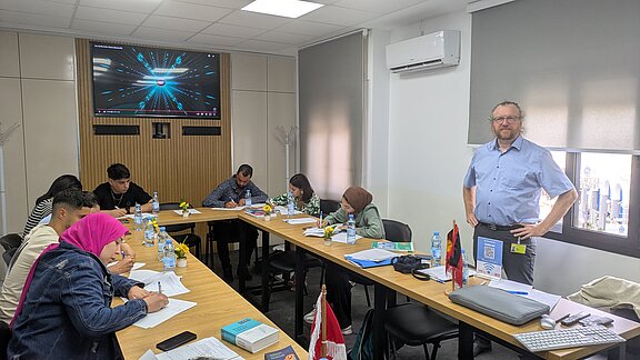 Eine Gruppe von vier Personen sitzt an einem Tisch und schreibt Notizen, während ein Mann im Hintergrund steht. Auf einem Bildschirm im Raum sind visuelle Elemente zu sehen. Es sind Wasserflaschen und kleine Pflanzen auf dem Tisch platziert.