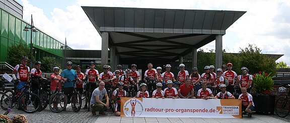 Gruppe von Radfahrerinnen und Radfahrern in roten Trikots posiert mit Fahrrädern vor einem Gebäude. Vorne ein Banner „radtour-pro-organspende.de“ und „TransDia Sport“. Gruppenfoto einer Benefizradtour zur Unterstützung der Organspende.