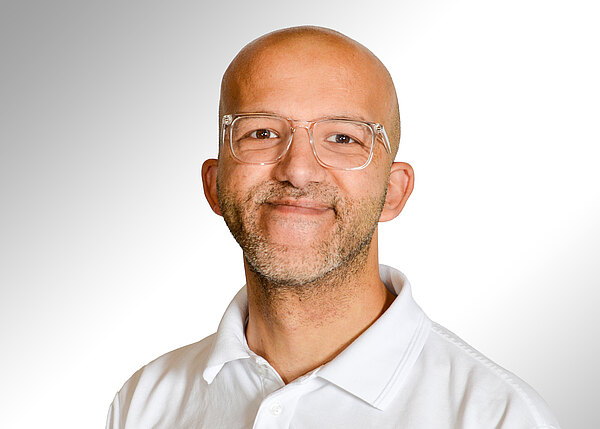 Porträt eines lächelnden, glatzköpfigen Mannes mit kurzer Bartstoppel und transparenter, eckiger Brille; er trägt ein weißes Poloshirt vor neutralem, hellgrauem Hintergrund.