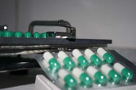 Eine Nahaufnahme zeigt eine Packung Kapseln mit grünen und weißen Farben. Im Hintergrund ist ein Teil eines Geräts zu sehen, das vermutlich zur Dosierung der Kapseln dient. Die Kapseln sind ordentlich in einer Reihenfolge angeordnet.