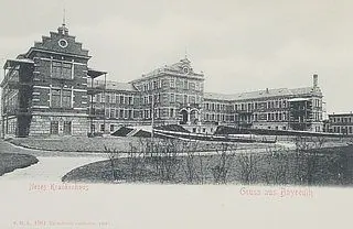 Das Bild zeigt ein historisches Krankenhausgebäude in Bayern. Es ist groß und beeindruckend, mit mehreren Etagen und einem klaren architektonischen Stil. Vor dem Gebäude erstreckt sich eine grüne Fläche, die einladend wirkt.