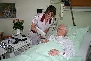 Eine Krankenschwester steht neben einem Bett, in dem eine ältere Patientin liegt. Die Krankenschwester hält ein Dokument in der Hand und spricht mit der Patientin. Auf dem Nachttisch stehen eine Pflanze und medizinische Utensilien.