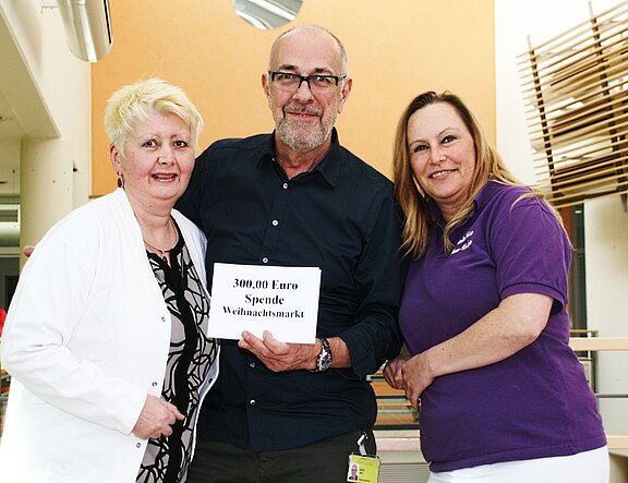 Drei Erwachsene posieren in einem Gebäude; die Person in der Mitte hält ein Schild mit der Aufschrift: „300,00 Euro Spende Weihnachtsmarkt“. Die Gruppe lächelt und präsentiert die Spende.