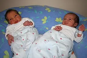 Zwei Neugeborene, eingehüllt in Decken, liegen nebeneinander. Das erste Baby hat braune Haare und schaut zur Seite, während das zweite Baby, mit dunklerem Haar, die Hand hebt. Beide tragen einfache, weiße Kleidung. Die Umgebung ist in sanften Farben gehalten.