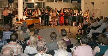 Eine Chorgemeinschaft steht auf einer Bühne und singt, während ein Pianist am Klavier begleitet. Das Publikum, bestehend aus älteren Menschen, sitzt in Reihen und hört aufmerksam zu. Die Atmosphäre ist festlich und musikalisch.