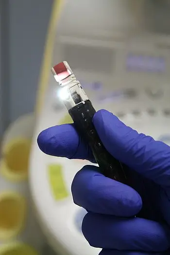 Eine Hand mit blauer Gummihandschuh hält ein medizinisches Gerät. Das Gerät hat eine rote Spitze und leuchtet. Im Hintergrund ist ein medizinisches Gerät mit Displays und Steuerungselementen zu erkennen.