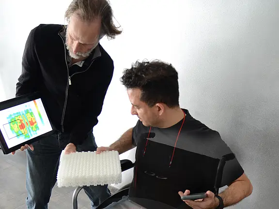 Zwei Personen prüfen die Sitzposition eines Rollstuhlfahrers. Der Stehende zeigt auf einem Tablet eine Druckverteilungs-Heatmap und hält ein geriffeltes Sitzkissen. Ziel ist die Anpassung der Sitzunterlage zur Druckentlastung und Komfortverbesserung.