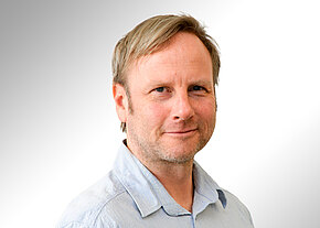 Dr. rer. pol. Reiner Hofmann Porträt eines freundlich blickenden Mannes mittleren Alters mit hellblonden Haaren und leichten Bartstoppeln, in hellblauem Hemd, vor neutralem, hellgrau-weißem Hintergrund.