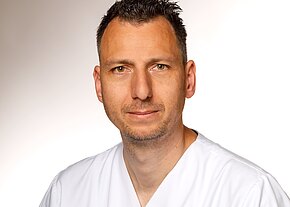 Porträt eines Mannes mittleren Alters in weißer medizinischer Kleidung vor neutralem Hintergrund. Er blickt ruhig in die Kamera.