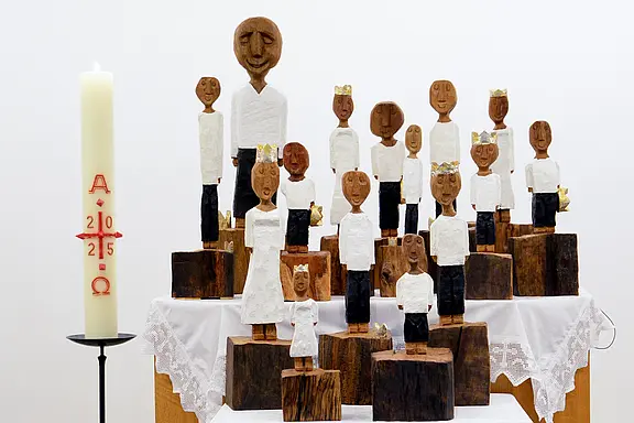 Mehrere geschnitzte Holzfiguren mit weißen Oberteilen stehen auf gestuften Holzblöcken wie eine Gemeinde; einige tragen goldene Krönchen. Links daneben eine große Osterkerze mit Alpha und Omega und der Jahreszahl 2025. Heller, sakraler Aufbau.
