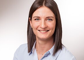 Christin Strobel Porträtfoto einer lächelnden Frau mit schulterlangem, glattem, dunkelbraunem Haar, leichter Goldkette und hellblauer Bluse. Sie blickt direkt in die Kamera vor neutralem, hellem Hintergrund.