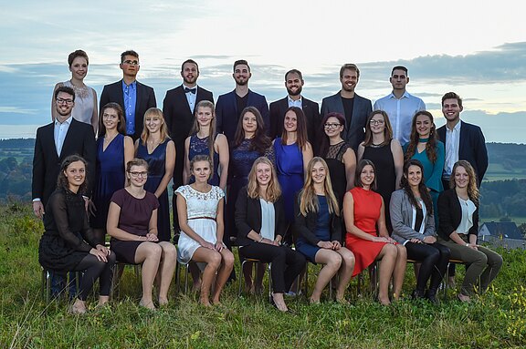 Eine Gruppe von 27 Personen steht und sitzt in zwei Reihen auf einer Wiese. Sie tragen formelle Kleidung und lächeln in die Kamera. Im Hintergrund ist eine malerische Landschaft mit sanften Hügeln und einem bewölkten Himmel zu sehen.