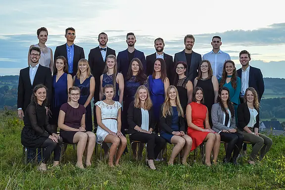 Eine Gruppe von 27 Personen steht und sitzt in zwei Reihen auf einer Wiese. Sie tragen formelle Kleidung und lächeln in die Kamera. Im Hintergrund ist eine malerische Landschaft mit sanften Hügeln und einem bewölkten Himmel zu sehen.