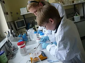 Zwei Personen in Laborkitteln arbeiten in einem Labor. Eine Person, ein Kind, misst mit einem Löffel eine Substanz in ein Gefäß, während die andere Person, eine Jugendliche, ihm zuschaut und Anweisungen gibt. Мeine Arbeitsumgebung enthält verschiedene chemische Behälter.