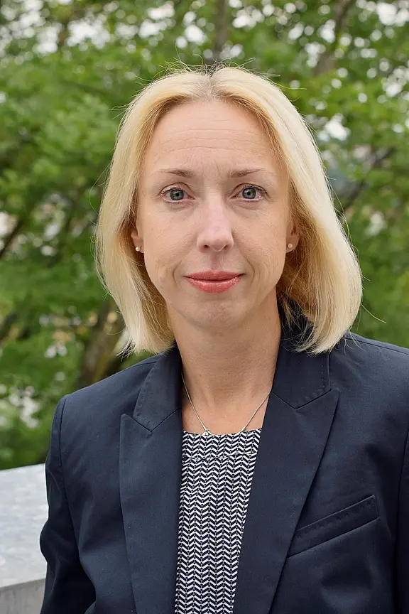 Porträt einer blonden Frau mit kurzen Haaren, die einen blauen Blazer und ein gestreiftes Oberteil trägt. Sie steht im Freien vor einer grünen Umgebung und schaut direkt in die Kamera.
