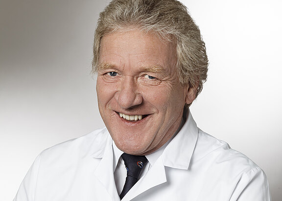 Porträt eines lächelnden Mannes im weißen Kittel und dunkler Krawatte vor neutralem Hintergrund; Kopf-und-Schulter-Aufnahme mit freundlichem, professionellem Ausdruck.