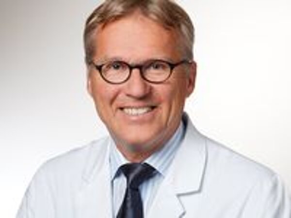 Ein lächelnder Arzt in weißem Kittel mit einer hellblauen Hemd und einer dunklen Krawatte. Er trägt runde Brille und hat kurzes, blondes Haar. Der Hintergrund ist hell und schlicht, um den Fokus auf den Arzt zu legen.