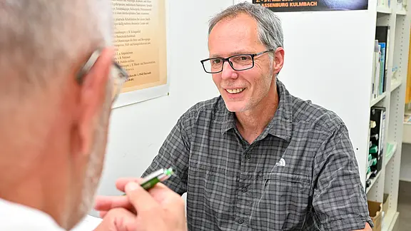 Zwei Männer sitzen sich in einem Büro gegenüber. Der im Vordergrund hält einen Stift, der andere, mit Brille und kariertem Hemd, lächelt und hört zu. Gesprächssituation, vermutlich Beratung oder Interview am Schreibtisch.