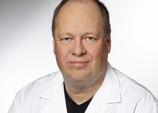 Porträt eines mittelalten Mannes in weißem Kittel vor neutralem Hintergrund, der direkt in die Kamera blickt, mit neutralem Ausdruck.