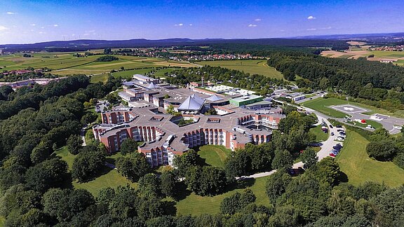 Luftaufnahme eines großen, ringförmig angelegten Klinik- oder Campuskomplexes in grüner Landschaft. Mehrere flache Gebäude mit Innenhöfen und eine zentrale Halle mit spitzem Dach, umgeben von Bäumen, Zufahrtsstraßen und Parkplätzen; dahinter Felder, Wälder und Hügel unter blauem Himmel.