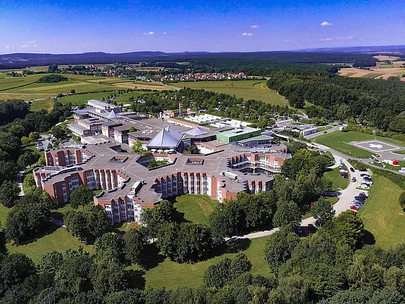 Luftaufnahme eines großen, ringförmig angelegten Klinik- oder Campuskomplexes in grüner Landschaft. Mehrere flache Gebäude mit Innenhöfen und eine zentrale Halle mit spitzem Dach, umgeben von Bäumen, Zufahrtsstraßen und Parkplätzen; dahinter Felder, Wälder und Hügel unter blauem Himmel.