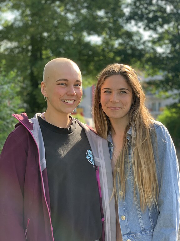 Zwei lächelnde Personen stehen eng nebeneinander im Freien. Links Person mit rasiertem Kopf, dunklem Shirt und Jacke; rechts Person mit langen, hellbraunen Haaren und Jeansjacke. Weiches Sonnenlicht, unscharfer grüner Hintergrund.