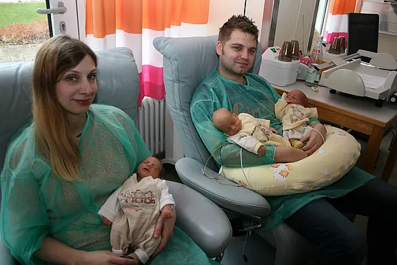 Zwei Erwachsene in Krankenhauskleidung sitzen in Sesseln und halten Neugeborene; der Mann trägt zwei Babys auf einem Stillkissen, die Frau hält ein Baby. Szene wirkt wie frischgebackene Eltern mit Drillingen im Krankenhaus.