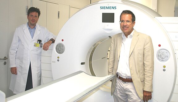 Zwei Männer stehen neben einem großen Siemens-Computertomographen in einem Klinikraum. Einer trägt einen weißen Arztkittel, der andere einen hellen Anzug. Das Gerät mit runder Öffnung und Liege ist bereit für eine Untersuchung.