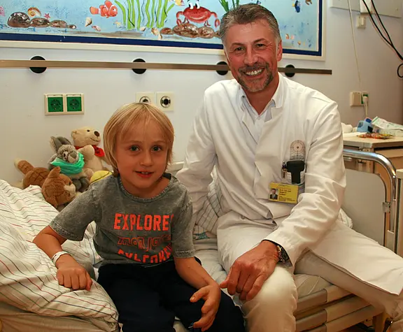 Ein lächelnder Arzt im weißen Kittel sitzt neben einem Jungen auf einem Krankenhausbett. Der Junge trägt ein T-Shirt, wirkt ruhig und hat ein Armband am Handgelenk. Beide schauen in die Kamera; die Szene vermittelt freundliche, vertrauensvolle Betreuung in einer Kinderklinik.