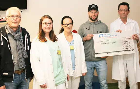 Fünf Personen stehen in einem Krankenhausraum. Drei tragen weiße Kittel. Eine Person in Freizeitkleidung hält einen großen symbolischen Scheck; daneben ein Arzt. Szene einer Spendenübergabe an medizinisches Personal.