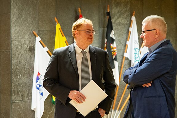 Zwei Männer im Anzug sprechen miteinander in einem Innenraum. Der Linke hält ein Dokument in der Hand. Hinter ihnen stehen mehrere Flaggen auf Ständern. Die Szene wirkt wie ein offizielles Treffen oder Empfang.