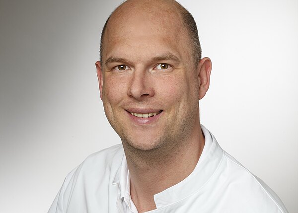 Porträtfoto: Ein lächelnder, glatzköpfiger Mann in weißem Kittel blickt direkt in die Kamera. Neutraler Hintergrund, sachlich-professionelle Darstellung.