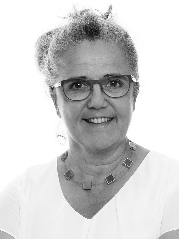 Schwarzweiß-Porträt einer älteren Frau mit grauen, hochgesteckten Haaren und Brille. Sie lächelt freundlich in die Kamera, trägt ein helles Oberteil und eine Kette mit quadratischen Elementen. Neutraler Hintergrund.