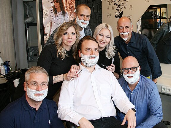 Gruppe in einem Friseursalon: Mehrere Männer sitzen und stehen mit Rasierschaum im Gesicht, zwei Stylistinnen halten Rasiermesser bereit. Alle lächeln in die Kamera; die Szene wirkt wie eine gemeinsame, humorvolle Rasuraktion.