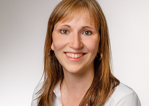 Porträt einer lächelnden Frau mit langen, braunen Haaren, die einen weißen Hemdkragen trägt. Der Hintergrund ist hell und neutral. Ihr Gesichtsausdruck ist freundlich und einladend.