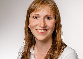 Porträt einer lächelnden Frau mit langen, braunen Haaren, die einen weißen Hemdkragen trägt. Der Hintergrund ist hell und neutral. Ihr Gesichtsausdruck ist freundlich und einladend.