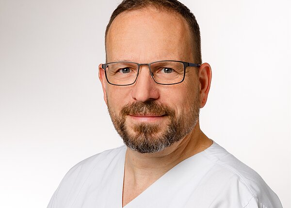 Porträt eines Mannes mit Brille und einem freundlichen Lächeln, der einen weißen Arztkittel trägt. Der Hintergrund ist neutral und hell. Die Person strahlt Kompetenz und Zutrauen aus.