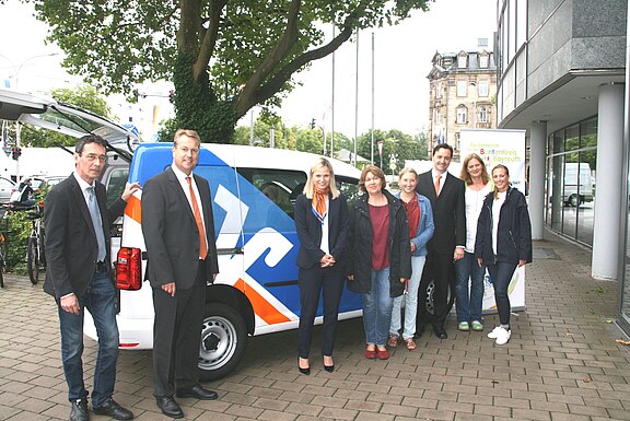 Gruppenfoto vor einem Firmenfahrzeug. Sieben Erwachsene stehen neben einem weiß-blauen Lieferwagen mit großem Logo, posieren lächelnd auf dem Gehweg vor einem Bürogebäude. Anlass wirkt wie Fahrzeugübergabe oder Präsentation.