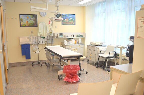 Helles Untersuchungszimmer: zentrale Untersuchungsliege mit Überkopf-Lampe, an der Wand medizinische Anschlüsse und Geräte; seitlich Schränke, Arbeitsfläche und Stühle; roter Abfallbehälter; großes Fenster mit Vorhängen.