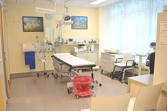 Helles Untersuchungszimmer: zentrale Untersuchungsliege mit Überkopf-Lampe, an der Wand medizinische Anschlüsse und Geräte; seitlich Schränke, Arbeitsfläche und Stühle; roter Abfallbehälter; großes Fenster mit Vorhängen.