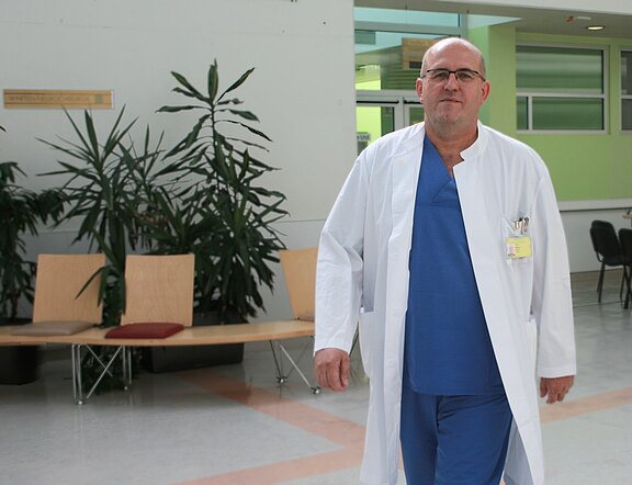 Ein Arzt in blauer OP-Kleidung mit weißem Kittel geht durch einen Krankenhausflur und schaut Richtung Kamera.