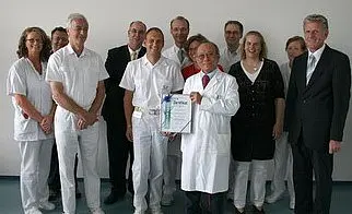Eine Gruppe von zehn Personen steht zusammen, darunter Ärzte und Pflegepersonal, und hält ein Zertifikat in den Händen. Alle lächeln, während sie für das Foto posieren. Der Moment zeigt Anerkennung und Teamarbeit in einem medizinischen Umfeld.