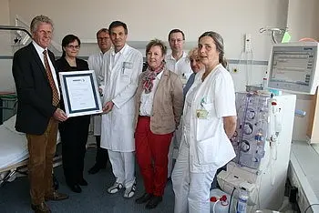 Eine Gruppe von sieben Personen in Krankenhausuniformen und Zivilkleidung steht zusammen in einem Raum. Sie halten ein Zertifikat in der Mitte der Gruppe, das eine Auszeichnung verkündet. Im Hintergrund ist ein medizinisches Gerät sichtbar.