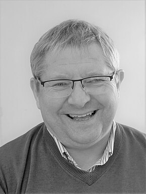 Schwarzweiß-Porträt eines freundlich lächelnden Mannes mittleren Alters. Er trägt eine Brille, kurzes helles Haar und schaut direkt in die Kamera. Kleidung: Pullover über einem gestreiften Hemd. Neutraler Hintergrund.